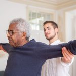 Idoso a fazer exercício de equilíbrio e força assistido por um cuidador em casa