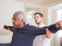 Idoso a fazer exercício de equilíbrio e força assistido por um cuidador em casa